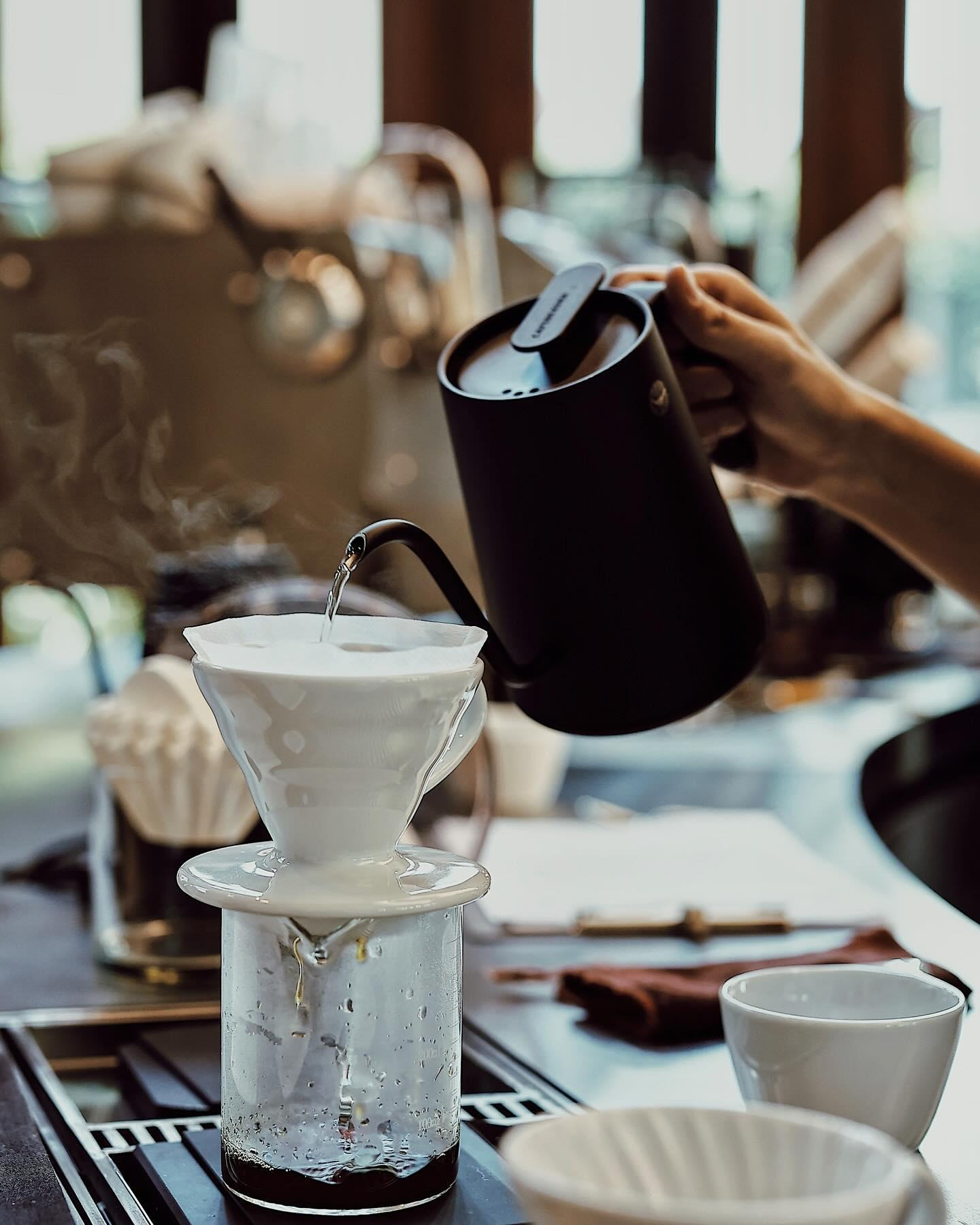 Close-up of a barista pouring hot water into a pour-over coffee dripper with steam rising.