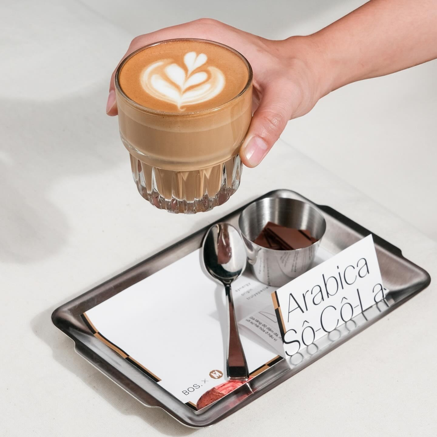Hand holding a latte art coffee over a tray with chocolate and a card.